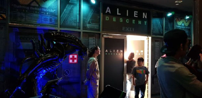 Photo of people entering the “Alien: Descent” building. A statue of an adult Xenomorph alien stands to the side of the entrance, with staff nearby.