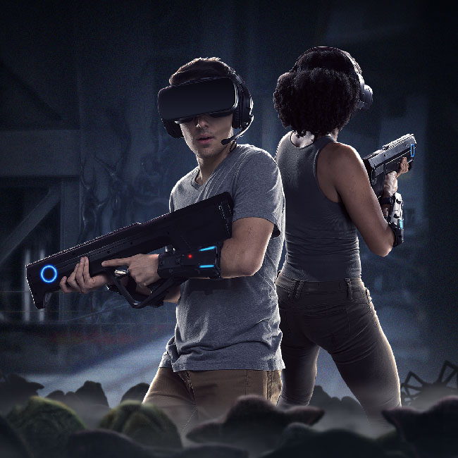 Promotional image of “Alien: Descent.” It shows two people wearing VR gear (visor headset, plastic gun, lightweight devices on their arms and legs) while in a dark shadowy area, surrounded by what seems to be alien eggs and at least one facehugger alien.