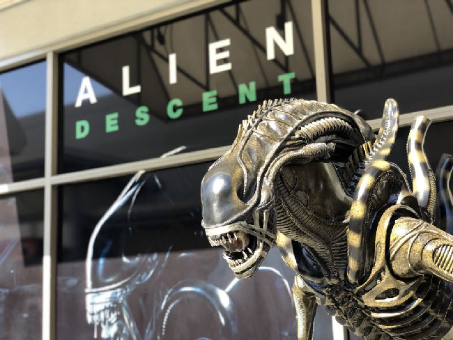 Photo of the exterior of the “Alien: Descent” building and Xenomorph alien statue. The building has a poster of “Alien: Descent” with an image of the Xenomoprh alien on it.