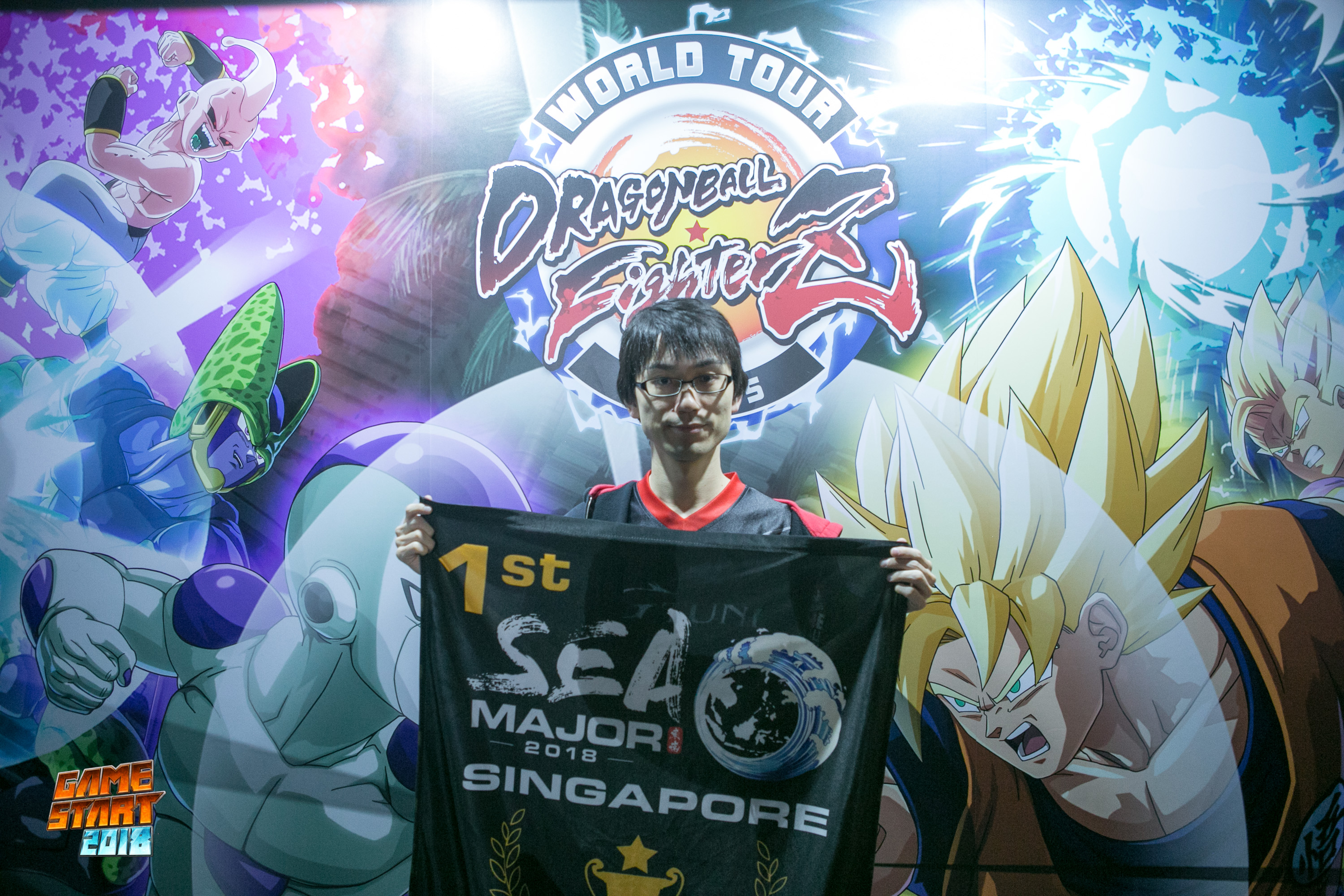 Photo of Ryota “Kazunoko” Inoue holding a first place South East Asia Major 2018 banner, and standing in front of a "Dragon Ball FighterZ" World Tour poster.